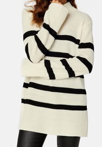 Crème sweater met zwarte horizontale strepen, ribbeltextuur, opstaande kraag en lange mouwen. Heeft verlengde mouwen die de handen bedekken.