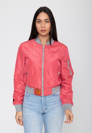 Woman wearing a pink bomber jacket with grey cuffs and collar, standing against a plain light background.