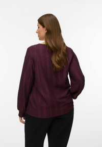 Femme aux longs cheveux bruns portant une blouse prune texturée à manches longues et un pantalon noir, debout de dos avec une main sur la hanche.