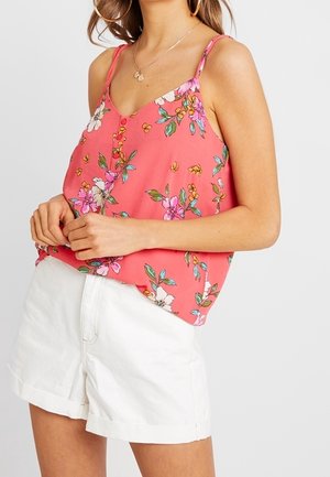 Kobieta ubrana w koralowo-różową kwiecistą bluzkę bez rękawów z guzikami, w zestawieniu z białymi szortami z wysokim stanem, stojąca na jednolitym tle.