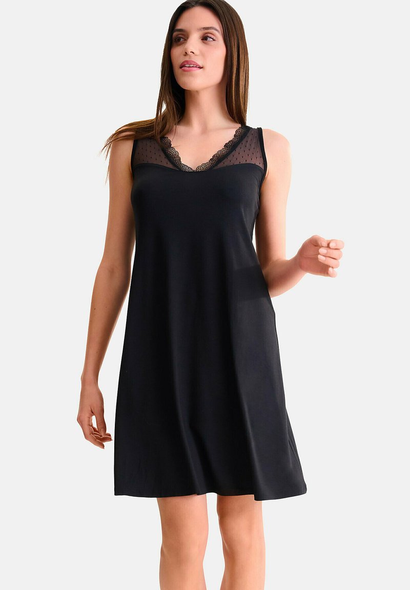 Femme portant une robe noire sans manches avec un col en polka dot transparent et une bordure en dentelle, se tenant devant un fond clair et uni.