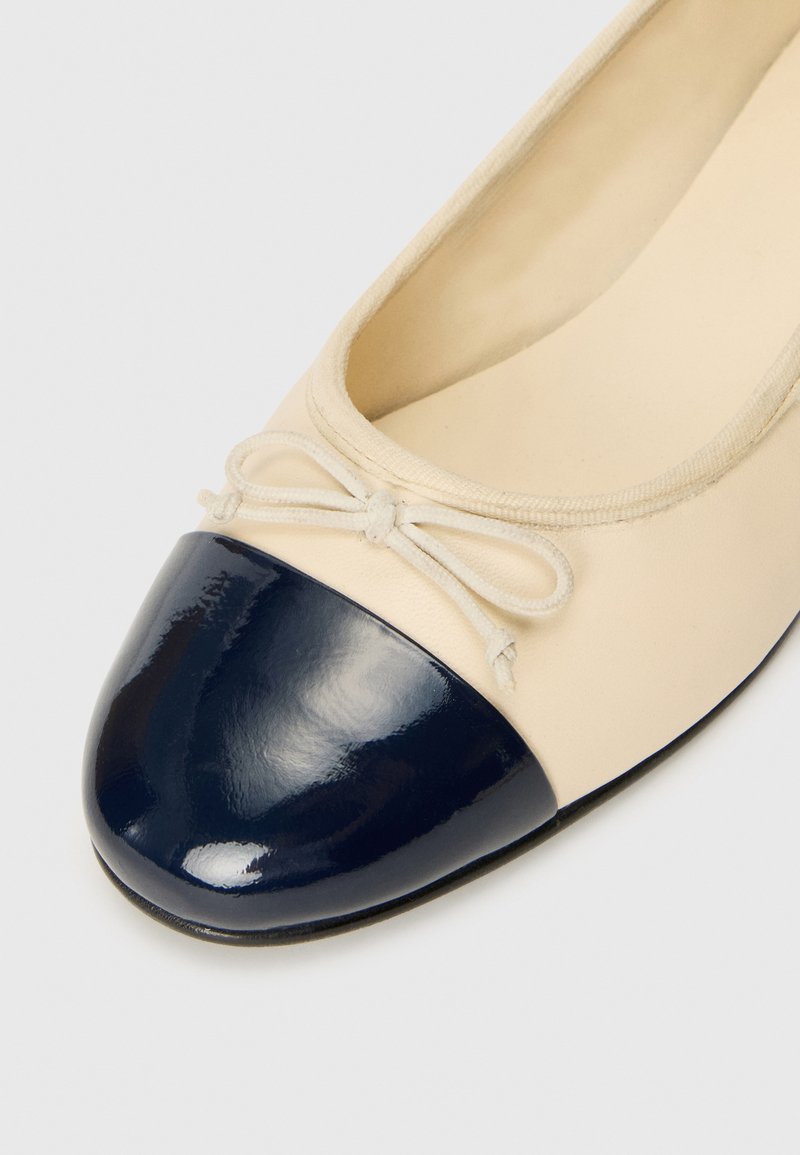 Ballerines beiges avec un bout en cuir verni bleu marine. Elles présentent un fin nœud à l'avant et un tissu lisse. Design classique.