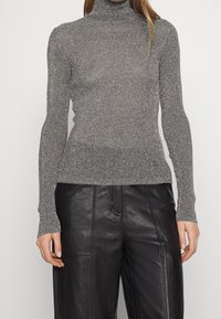 Femme portant un pull à col roulé argenté métallisé à manches longues et un pantalon en cuir noir, debout devant un fond blanc uni.