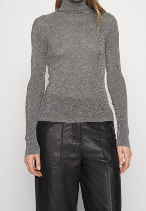 Femme portant un pull à col roulé argenté métallisé à manches longues et un pantalon en cuir noir, debout devant un fond blanc uni.