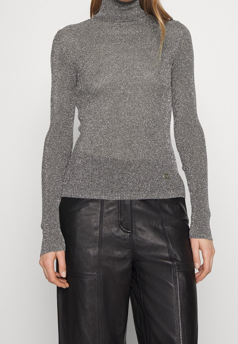 Femme portant un pull à col roulé argenté métallisé à manches longues et un pantalon en cuir noir, debout devant un fond blanc uni.