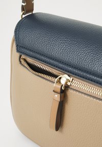 Brown leather crossbody bag with textured surface, navy top flap, gold zipper, and a tan strap. Features a small zippered pocket.