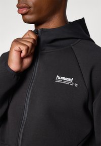 Uomo che indossa una felpa nera con cappuccio e cerniera, tirando la zip verso l'alto vicino al collo, mostrando il logo "hummel" sul petto.