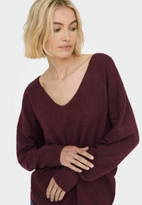 Ženska s kratkimi blond lasmi, oblečena v ohlapno burgundno pulover s globokim V-izrezom in dolgimi rokavi, gleda navzdol in si prilagaja rokav.