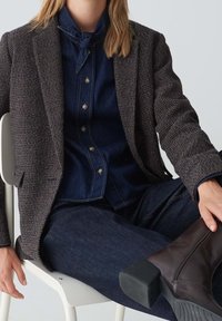 Personne portant un blazer à carreaux foncés par-dessus une chemise en denim nouée et un jean, assise sur une chaise blanche avec une jambe croisée montrant des bottes marron foncé.