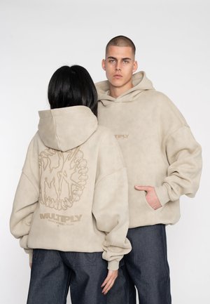 Las sudaderas beige de gran tamaño presentan un gran gráfico y logotipo en la parte posterior. Tejido suave con textura y puños acanalados, combinadas con jeans azul oscuro.