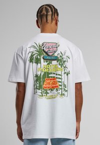 Hvid bomulds t-shirt med et farverigt grafisk tryk på ryggen, der inkluderer palmetræer, hotelbilleder og tekstdetaljer.