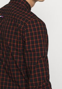 Hemd mit Knopfverschluss aus schwarzem Stoff mit einem rot-gelben Karomuster und einem umgeschlagenen Kragendetail auf der Rückseite.