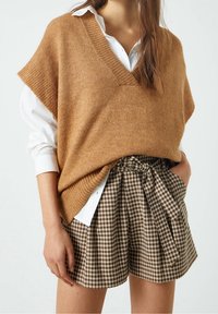 Top sin mangas de punto marrón con escote en V, usado sobre una camisa blanca con cuello, combinado con pantalones cortos de cuadros marrones de tiro alto con cintura ajustable.