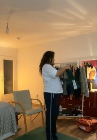 Pessoa vestindo uma camisa branca e calças pretas segura uma peça de roupa verde com três riscas brancas, rodeada por um cabide de roupas e mobília.