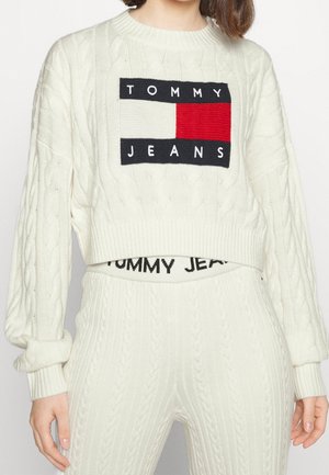 Persona con un conjunto de suéter de punto trenzado blanco y pantalones, con un logo de Tommy Jeans en negro, blanco y rojo en la parte delantera del suéter.