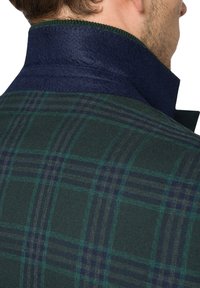 Marynarka w kratę w zielono-granatowym wzorze, z wysokim kołnierzem z ciemnoniebieskiego materiału i teksturowanym wełnianym wykończeniem przy kołnierzu.