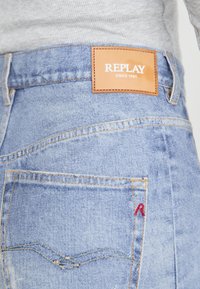 Svetlo modri jeans iz denima z usnjeno etiketo z napisom "REPLAY SINCE 1981," šivanim vzorcem na žepih in majhnim rdečim akcentom "R".