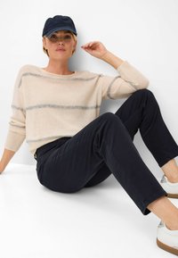 Un pull beige clair avec des rayures grises, un pantalon noir et une casquette marine. Les chaussures sont blanches avec des accents argentés. Assis contre un fond blanc.