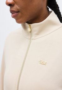 Záber zblízka na osobu v béžovej bunde na zips s vysokým golierom a zlatým logom EA7 na hrudi.