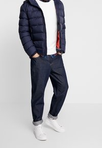 Marineblaue Puffjacke, weißer Pullover, kariertes Hemd, dunkle Jeans und weiße Sneaker. Die Jacke hat einen Reißverschluss und eine Kapuze. Aufgekrempelte Säume an den Jeans.