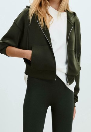Mujer con una sudadera con capucha verde oscuro con cremallera sobre una camiseta blanca y leggings negros, con una mano en el bolsillo de la sudadera.
