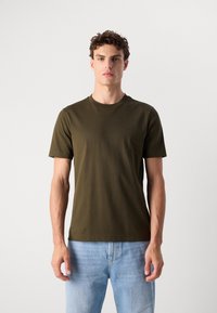 T-shirt en coton vert olive avec un col rond et des manches courtes. Présente un logo discret sur la poitrine. Assortie à un jean bleu clair.