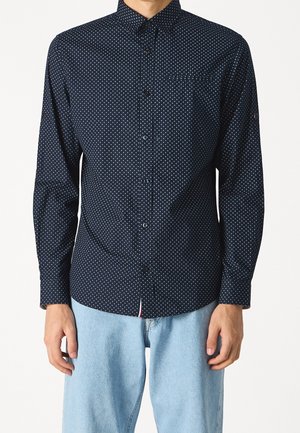 Muž v námořnicky modré košili s dlouhým rukávem a bílými puntíky a v světlých modrých džínách, stojící před bílým pozadím.