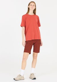 Punainen lyhythihainen t-paita yhdistettynä ruskeisiin shortseihin, joissa on useita taskuja. Mallilla on päällään vaaleanbeiget vaelluskengät ja valkoiset nilkkasukat.