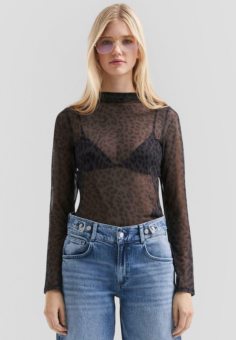 Schwarzes Mesh-Oberteil mit langen Ärmeln und Leopardenmuster, über einem schwarzen BH getragen, kombiniert mit hellen Denim-Jeans und goldfarbenen Sonnenbrillen.