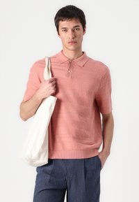 Polo majica kratkih rukava od teksturirane svijetloružičaste pletenine, s ovratnikom i zaklopkom na dva gumba. Model drži kremastu torbu.