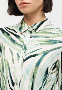 Γυναίκα που φοράει πουκάμισο με γιακά και κουμπιά, με πράσινες, μαύρες και μπλε αφηρημένες ρίγες ζέβρας σε λευκό ύφασμα.