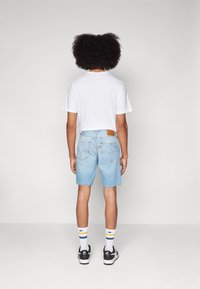 Henkilö, jolla on afro-hiustyyli, pukeutuneena valkoiseen t-paitaan, vaaleansinisiin farkkushortseihin, valkoisiin raidallisiin sukkiin ja musta-valkoisiin tennareihin, seisoo selin.