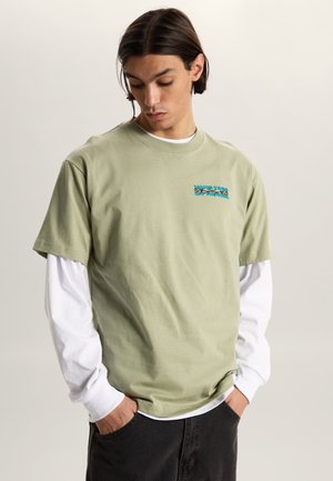 Joven con cabello oscuro que lleva una camiseta de manga larga blanca en capas bajo una camiseta gráfica Vans de color verde claro y pantalones oscuros.