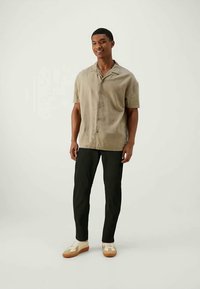 Homme portant une chemise beige à manches courtes, un pantalon noir et des baskets blanches, debout et souriant devant un fond clair et uni.
