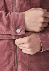 Mains ajustant le poignet d'une veste en velours côtelé rose avec un bouton-pression argenté et une fermeture éclair centrale.