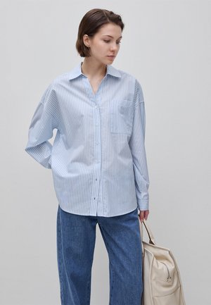 Frau in locker sitzendem blau-weiß gestreiftem Hemd mit Knöpfen und blauer Jeans, die eine beige Tasche hält und nach links unten schaut, vor schlichtem Hintergrund.