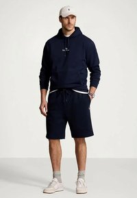 Άνδρας που φορά navy hoodie, ταιριαστά σορτς, γκρίζες κάλτσες, λευκά αθλητικά παπούτσια και λευκό καπέλο, στέκεται με το ένα χέρι στην τσέπη του σε επίπεδο φόντο.