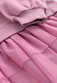 Gros plan d’un vêtement rose montrant un poignet côtelé et un tissu tulle transparent superposé.