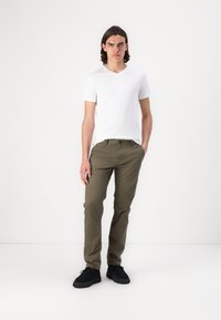 Valkoinen V-kaula-aukkoinen T-paita, oliivinvihreät slim-fit-housut, mustat kengät. Malli seisoo yksi käsi taskussa yksinkertaista valkoista taustaa vasten.