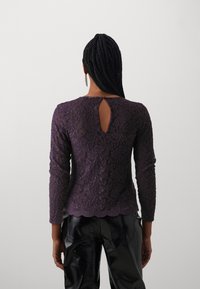 Lila Spitzen-Longsleeve-Oberteil mit gewelltem Saum und einer Schlüssel-Ausschnitt-Detail auf der Rückseite, kombiniert mit glänzenden schwarzen Hosen, die Textur und Design hervorhebt.