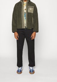 Zielona polarowa kurtka z przednim zamkiem, z kieszenią naszywaną, noszona na koszuli w kwiatowy wzór. Czarna spodnie i niebiesko-białe sneakersy dopełniają wygląd.