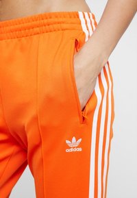 Orange idrottsbyxor med vita vertikala ränder och en sidficka. Har en broderad vit logotyp på vänstra låret. Slät tygtextur.