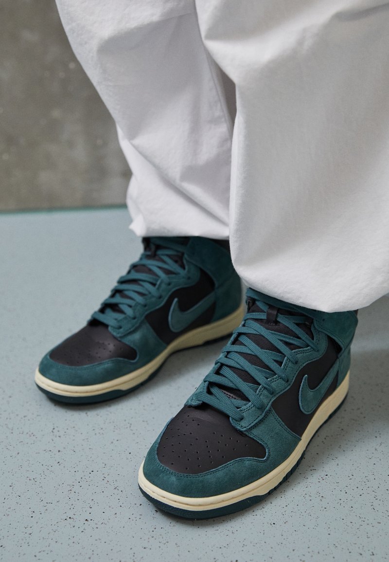 Korkea Nike-tennis, joissa on turkoosivihreää mokkaa ja mustaa nahkaa, rei'itetty varvasosa ja valkea kumipohja. Lankakuvio edessä.