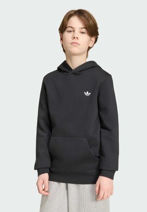 Junge Person mit braunen Haaren, die einen schwarzen Adidas-Hoodie mit Logo trägt und vor einem einfarbigen hellgrauen Hintergrund steht.