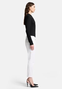 Maglietta nera a maniche lunghe, design aderente, abbinata a jeans bianchi slim-fit. Il modello indossa scarpe nere con tacco alto e punta affusolata.