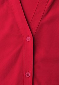 Gros plan sur le devant d’une chemise rouge avec deux boutons roses et des coutures visibles le long de la patte de boutonnage et du col.