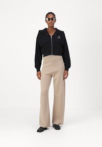 Felpa nera corta con zip, con logo. Abbinata a pantaloni beige a gamba larga. Modello che indossa scarpe nere e occhiali da sole scuri.