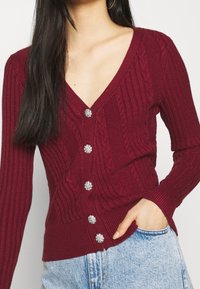 Burgundské pletené kardigan s výstřihem do V, žebrovanou strukturou a pěti lesklými knoflíky vpředu. Dlouhé rukávy a přiléhavý lem.