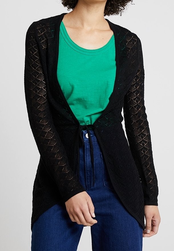Femme portant un cardigan noir en maille ouverte noué à la taille par-dessus une chemise verte et un jean bleu taille haute, cadré aux épaules et aux hanches.