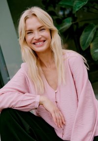 Blouse en tissu froissé rose clair avec un décolleté en V profond, dotée d'accents de boutons et de manches longues, associée à un pantalon noir.